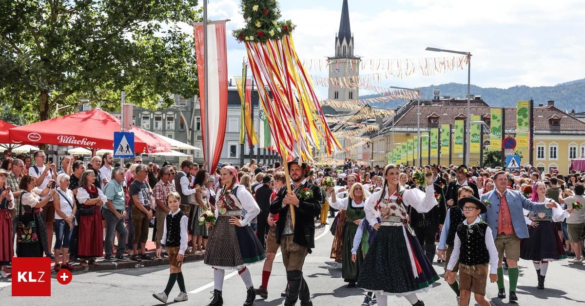 Ein festlicher Umzug mit Menschen in traditionellen Kostümen, die mit Blumen und Bannern durch die Straße ziehen. Im Hintergrund befinden sich Gebäude und ein Turm.