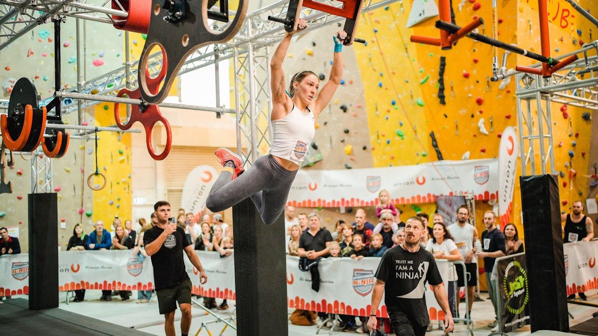 Eine Frau in Sportkleidung hängt kopfüber von einer Stange in einem Fitnessstudio. Zuschauer beobachten von hinter einer Barriere. Ein Mann macht ein Foto mit einem Handy.