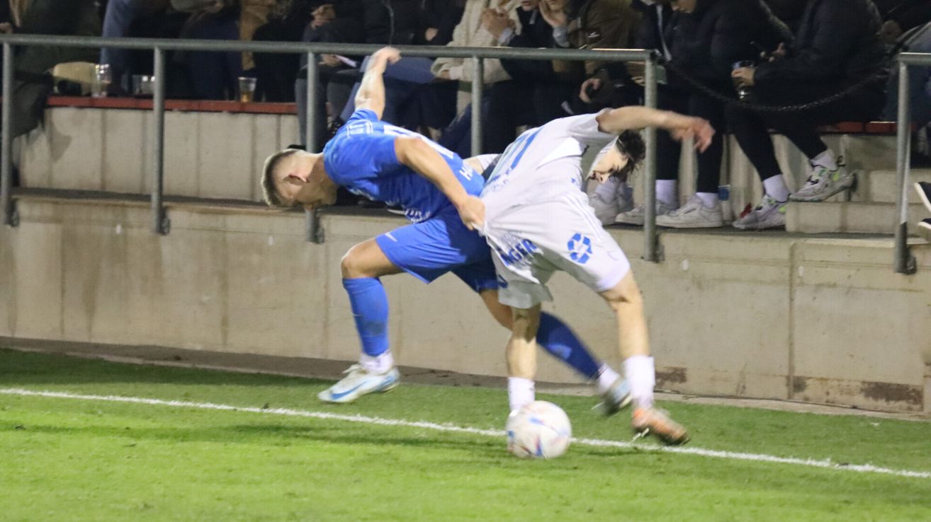 Two soccer players, one in blue and the other in white, are fighting for the ball on the field. Behind them, spectators are watching from the stands.