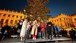 Eine Gruppe von Menschen steht auf einer Bühne vor einem Weihnachtsbaum und singt vor einem großen, beleuchteten Gebäude. Sie tragen Winterkleidung und der Bereich ist mit einem Publikum überfüllt.