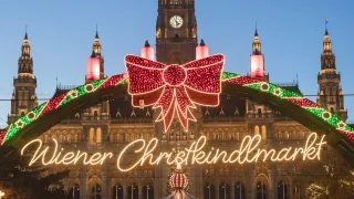 Beleuchteter Wiener Christkindlmarkt vor einem großen Gebäude mit Uhrturm, dekorativen Lichtern und einem Weihnachtsbaum.