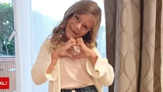 Eine Frau mit Brille und einem weißen Cardigan lächelt und formt mit ihren Händen ein Herz vor ihrer Brust.