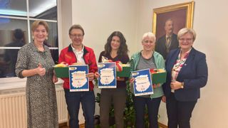 Drei Personen in einem Raum halten blaue Urkunden mit Nummern und einer grünen Geschenkbox. Hinter ihnen steht ein Porträt eines Mannes in Anzug.