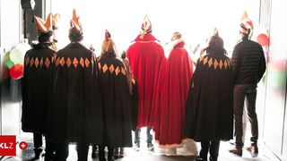 Eine Gruppe von Menschen in Kostümen, einige mit roten Umhängen und andere mit schwarzen Umhängen, stehen in einer Reihe und blicken in die gleiche Richtung. Sie nehmen wahrscheinlich an einer Parade teil.