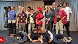 Eine Gruppe von Menschen in verschiedenen Kostümen, einschließlich Hüten und Kleidern, posiert für ein Foto auf der Bühne. Einige knien und andere stehen.