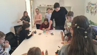 Eine Gruppe von Kindern sitzt um einen Tisch herum und beschäftigt sich mit einer Aktivität. Ein Kind konzentriert sich auf ein Handwerk, während andere zuschauen. Dahinter beobachtet ein Mann und einige weitere Kinder stehen.