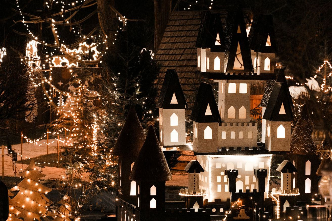 Ein Burgmodell ist mit Lichtern beleuchtet und neben einem mit Lichtern geschmückten Weihnachtsbaum platziert. Die Burg hat spitze Dächer und gewölbte Fenster. Die Lichter reflektieren von den Fenstern.