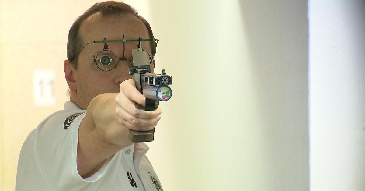 Ein Mann mit Brille und einer Pistole, er zielt auf ein Ziel, trägt ein weißes Hemd mit einem Logo auf der linken Brust.