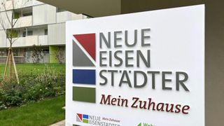 Ein Schild für Neue Eisenstadter Mein Zuhause steht vor einem modernen Gebäude mit Grünpflanzen. Das Schild zeigt rote, blaue, graue und grüne Farben.