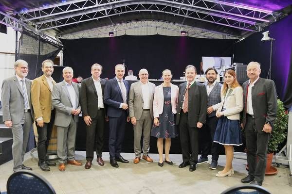 Eine Gruppe von Menschen in formeller Kleidung steht auf einer Bühne mit Hintergrund, möglicherweise für ein Foto.