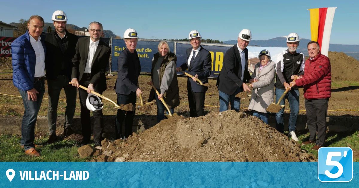 Mehrere Personen stehen in einem Feld, halten Schaufeln und tragen weiße Helme. Sie posieren für ein Foto. Dahinter ist ein Haufen Erde. Ein Banner befindet sich am Zaun.