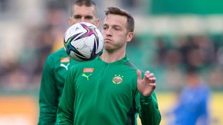 Ein Fußballspieler in einem grünen Trikot mit dem Wort Rapid Wien hält einen Fußball in der Luft. Ein anderer Spieler steht hinter ihm.