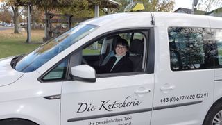 Eine Frau mit Brille sitzt in einem weißen Taxi-Van mit den Worten Die Kutscherin an der Seite, geparkt in einem Außenbereich.