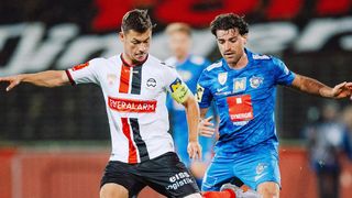 Zwei Fußballer in Aktion, einer in Weiß und Rot und der andere in Blau, kämpfen während eines Spiels um den Ball.