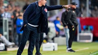 Ein Trainer zeigt mit der Hand Richtung Spielfeld, trägt eine blaue Jacke und eine Uhr. Hinter ihm stehen andere Personen, darunter ein weiterer Trainer, an der Seitenlinie.