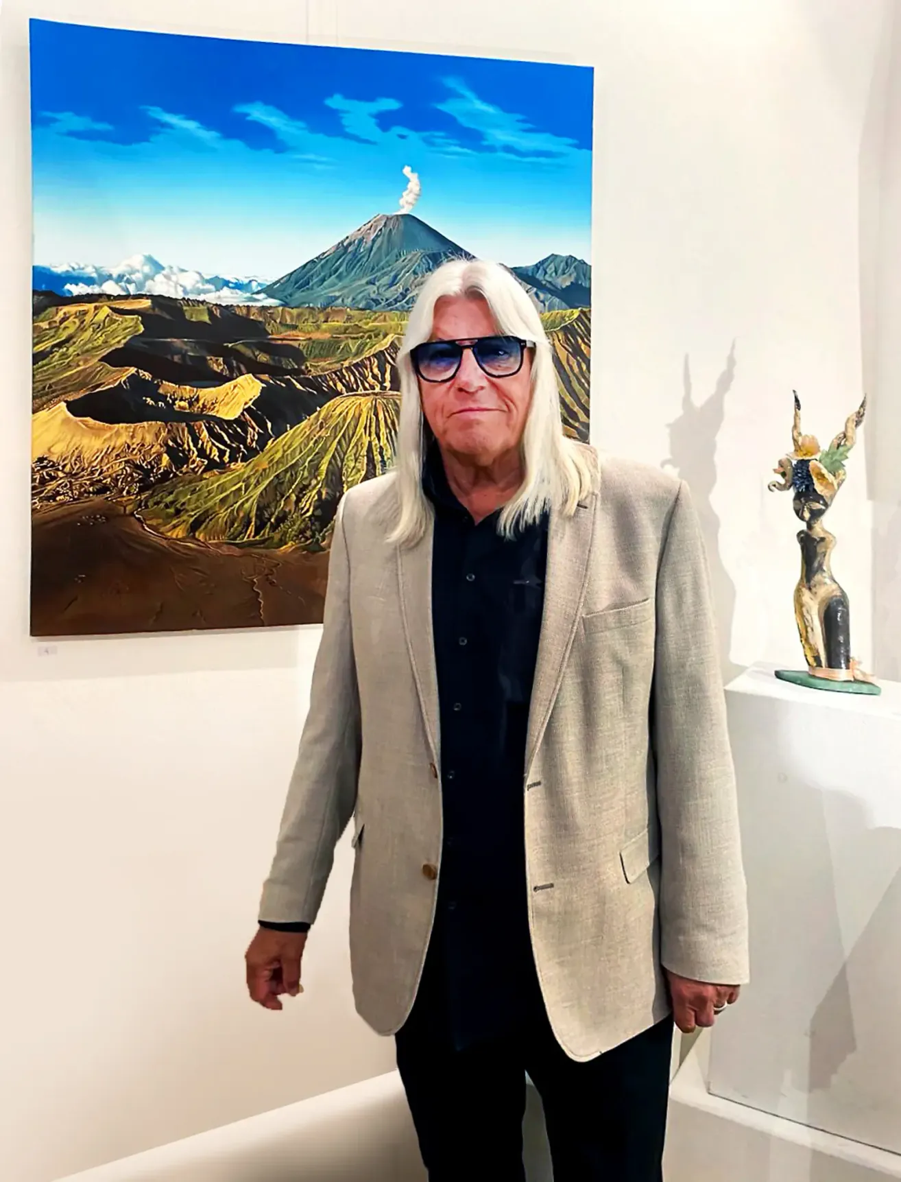 Ein Mann mit weißen Haaren und einer Brille steht vor einem großen Gemälde, das eine Berglandschaft mit einem Vulkan im Hintergrund darstellt.