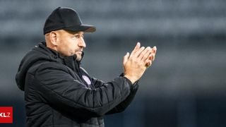 Ein Mann in einer schwarzen Jacke und Mütze klatscht, während er auf einem Feld steht und ernst aussieht.