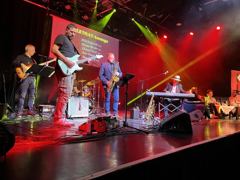 Eine Band tritt auf der Bühne auf, unter einem großen Banner, mit einem Gitarristen, Saxophonisten und Keyboarder. Rote und grüne Scheinwerfer beleuchten die Szene, und im Hintergrund befinden sich ein Schlagzeug und ein Mikrofonstativ.