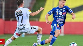 Zwei Fußballspieler rennen auf dem Feld, einer trägt ein weißes Trikot mit der Nummer 11 und der andere ein blaues Trikot mit verschiedenen Werbeaufdrucken.
