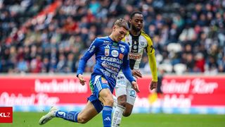 Zwei Fußballspieler in Aktion, einer in Blau und einer in Schwarz und Gelb. Der Spieler in Blau dribbelt den Ball, während der Gegner versucht, ihn zu tackeln. Der Hintergrund zeigt ein Stadion, das mit Zuschauern gefüllt ist.