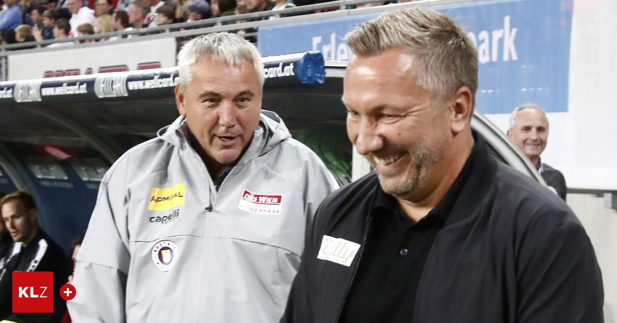 Zwei Männer stehen nebeneinander, lächeln und schauen in die Kamera. Der Mann auf der linken Seite trägt eine graue Jacke mit einem gelben Logo und dem Wort 'Capelli' darauf. Der Mann auf der rechten Seite trägt ein schwarzes Hemd. Dahinter sind verschwommene Bilder von Menschen zu sehen, die auf den Tribünen sitzen.