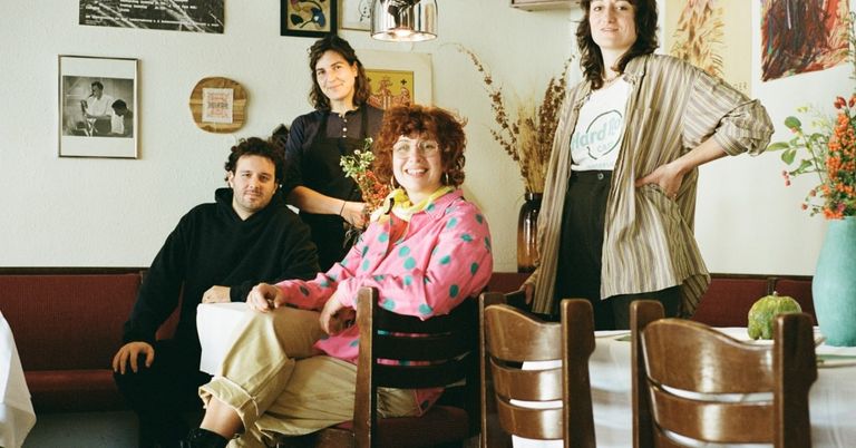 Four people sit at a table in a room. A woman in a pink polka-dot jacket smiles at the camera. Behind her, a woman in a black apron holds flowers. On the right, a woman in a striped shirt poses with a hand on her hip.