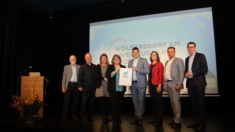 Eine Gruppe von Menschen steht auf der Bühne bei einer Preisverleihung und hält ein Zertifikat. Eine Leinwand hinter ihnen zeigt ein Logo und Text über 'Workers' Village AM Future'.