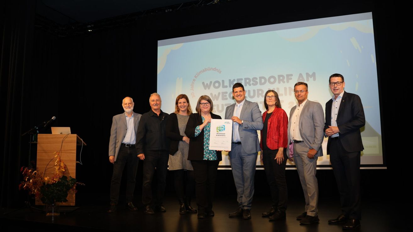 Eine Gruppe von Menschen steht auf der Bühne bei einer Preisverleihung und hält ein Zertifikat. Eine Leinwand hinter ihnen zeigt ein Logo und Text über 'Workers' Village AM Future'.