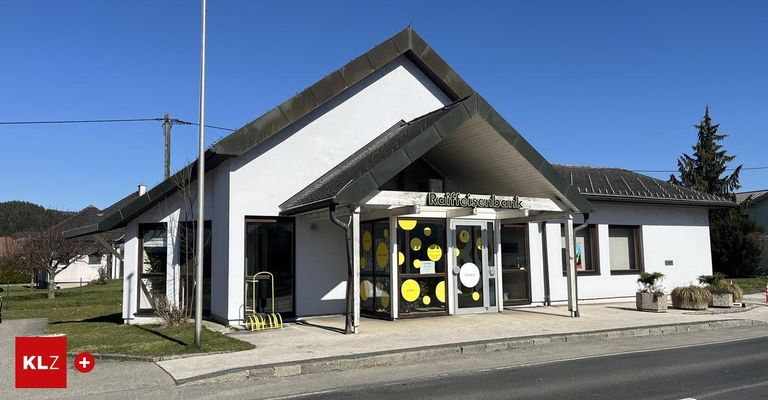 Ein weißes Gebäude mit schwarzem Dach und gelben Kreisen auf den Fenstern. Es ist eine Raiffeisenbank-Filiale. Links steht ein gelber Fahrradständer.