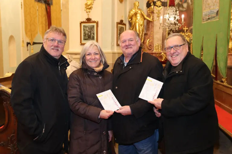 Four people stand in front of a golden statue, smiling and holding certificates. They wear winter coats and are likely in a church. Behind them is a chandelier and a wall with picture frames.