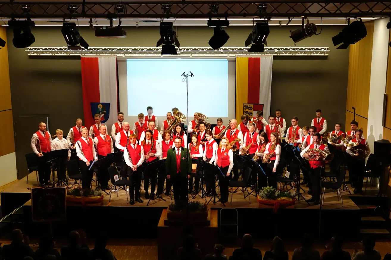 Ein Orchester spielt auf der Bühne, mit verschiedenen Instrumenten. Sie sind in roten Westen gekleidet. Eine große Leinwand befindet sich hinter ihnen.