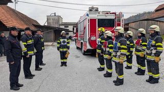 Eine Gruppe von Feuerwehrleuten steht in einer Reihe, mit einem Feuerwehrauto im Hintergrund, möglicherweise in Vorbereitung auf eine Übung oder Inspektion.