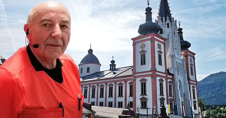 An older man with a red shirt stands in front of a church with domes and spires on a cloudy day.
