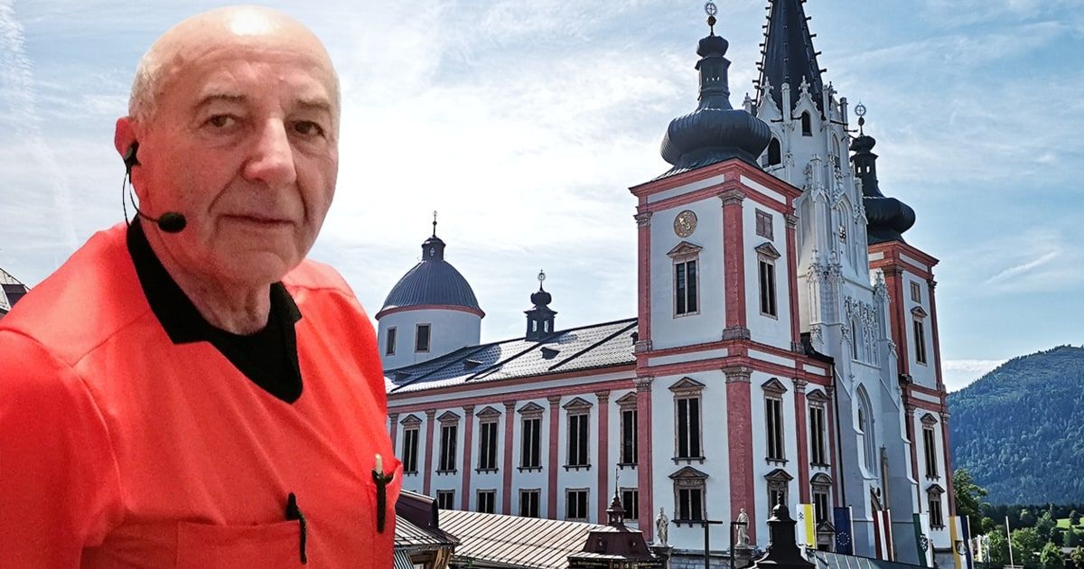 An older man with a red shirt stands in front of a church with domes and spires on a cloudy day.