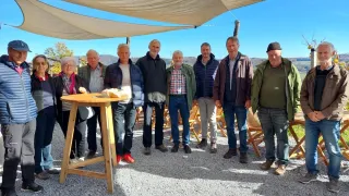 Eine Gruppe von Menschen steht an einem sonnigen Tag unter einem Vordach. Sie sind in verschiedenen Jacken und Hosen gekleidet, einige tragen Brillen. Ein Holztisch hält ein Brot. Die Umgebung ist von Holzstühlen und einer Kiesoberfläche umgeben.