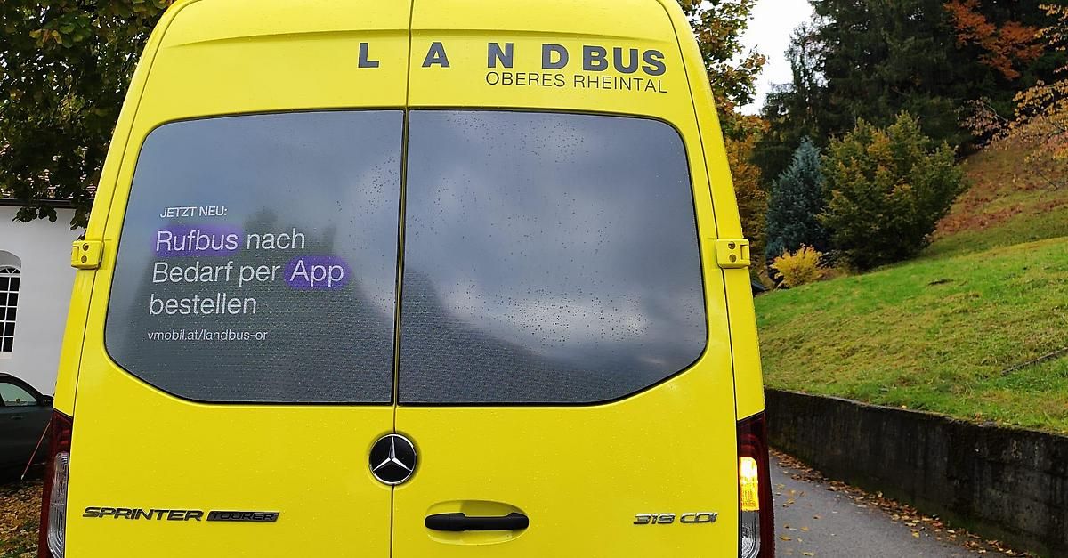 Ein gelber Bus mit 'LAND BUS OBERES RHEINTAL' an der Seite. Die Heckscheibe zeigt eine Reflexion von Bäumen und einem grasigen Hügel. Eine Werbung für das Aufrufen des Busses per App ist auf der Scheibe.