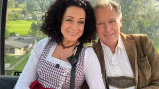 Ein lächelndes Paar in traditioneller Kleidung posiert für ein Foto aus einer Seilbahn, mit Blick auf ein malerisches Tal und Berge.