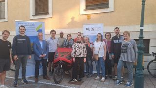 Eine Gruppe von Menschen steht vor einem Gebäude mit einem roten Fahrrad davor. Sie lächeln und posieren für ein Foto. Das Fahrrad hat eine rote Abdeckung.