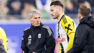 Ein Fußballtrainer in einer schwarzen Jacke unterhält sich mit einem Spieler in einem gelben und weißen Trikot mit der Nummer 11 darauf.