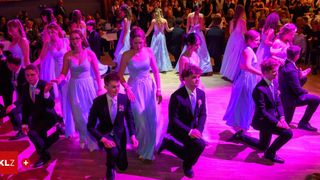 Mehrere junge Menschen in lila Kleidern und Anzügen tanzen auf einer Bühne mit lila Beleuchtung vor einem Publikum.