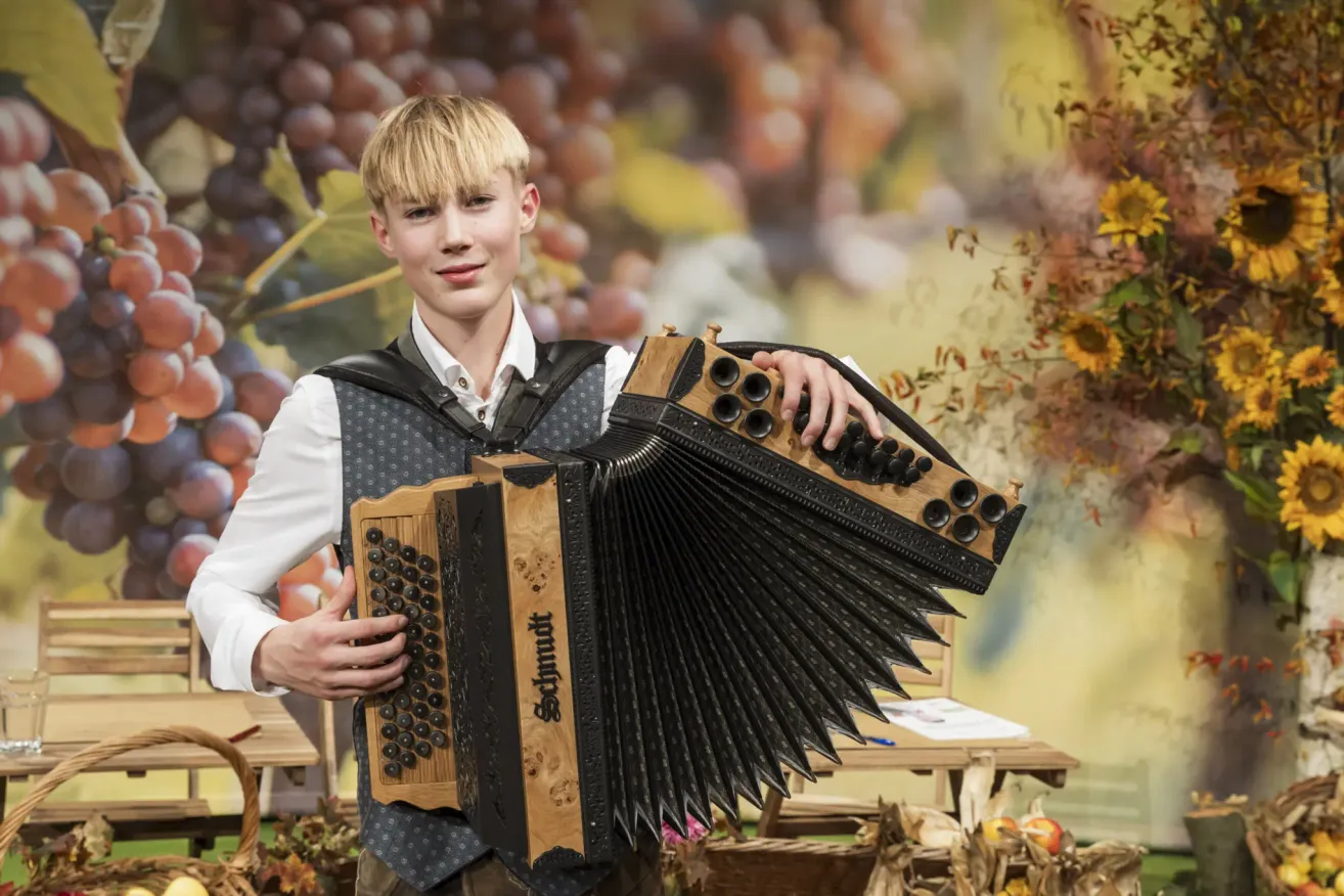 Ein junger Mann mit blondem Haar hält ein Akkordeon vor einem Hintergrund aus Trauben und Sonnenblumen. Er ist in einem weißen Hemd und einer Weste gekleidet.