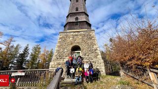 Eine Gruppe von Wanderern, einige mit Hunden, posiert für ein Foto vor einem Steinturm mit einem Kirchturm, umgeben von Bäumen.