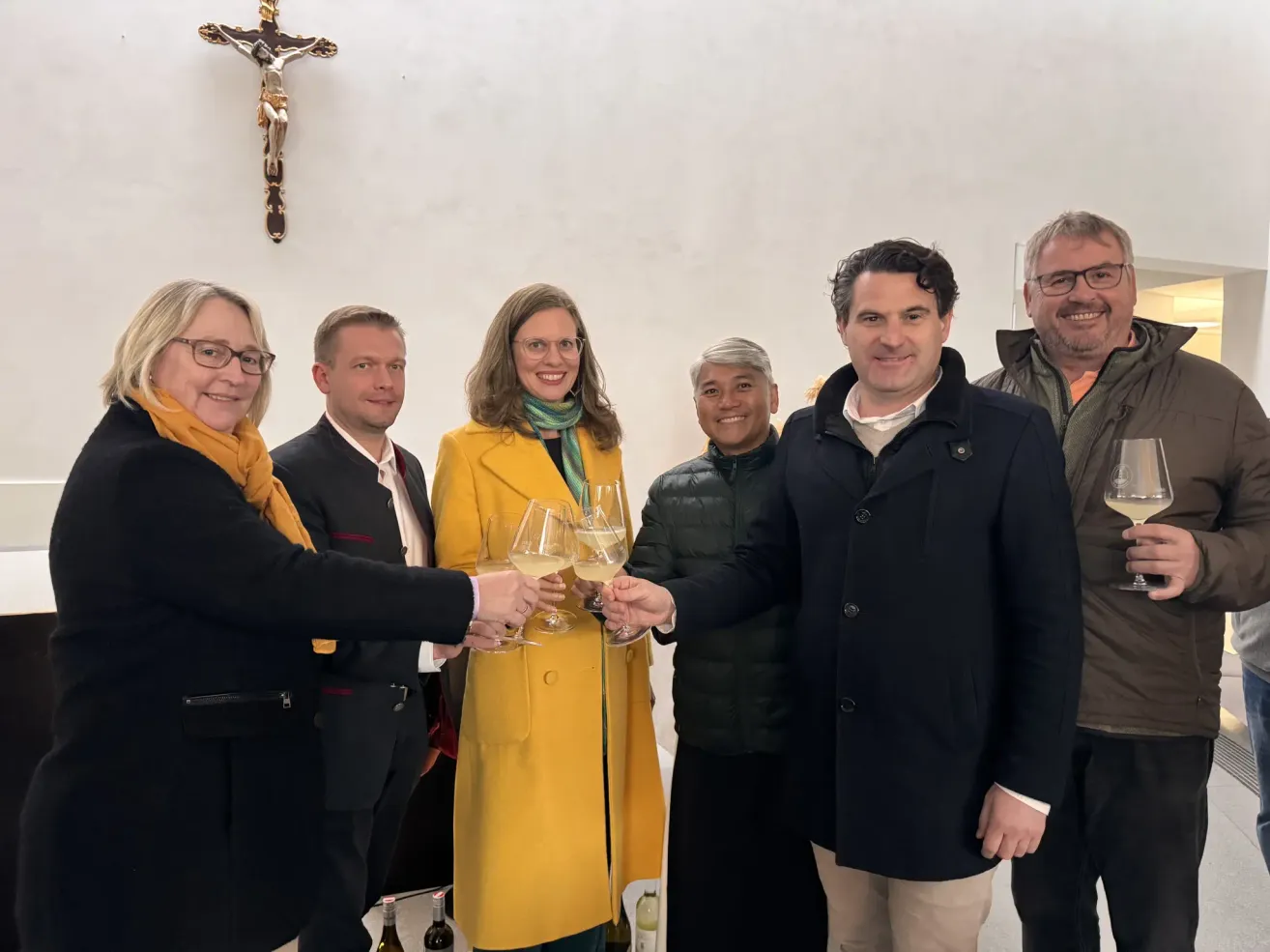 Eine Gruppe von sechs Personen, die formell gekleidet sind, lächeln und halten Weingläser in der Hand, wahrscheinlich bei einer Feier. Sie stehen vor einer weißen Wand mit einem Kreuz darauf.
