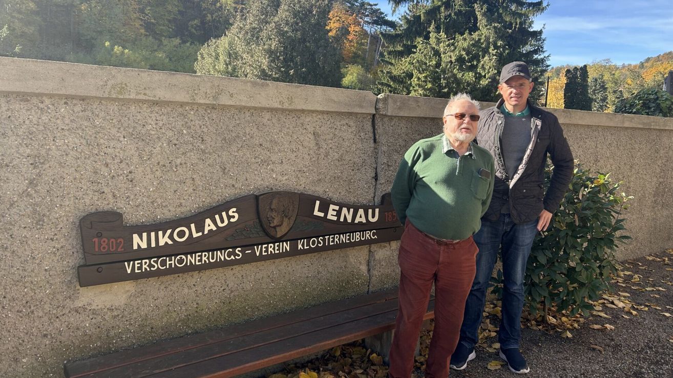 Zwei Männer stehen vor einer Steinmauer, einer trägt einen grünen Pullover und rote Hosen, der andere eine Jacke und Jeans. Dahinter befindet sich ein Schild mit der Aufschrift 'Nikolaus Lenoau 1835'.