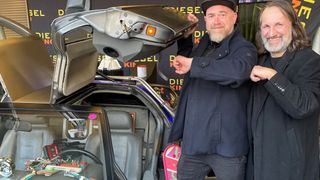 Zwei Männer stehen neben einem DeLorean-Auto mit geöffneter Motorhaube und zeigen das Innere des Autos und einen offenen Kofferraum mit einem Skateboard in der Nähe.