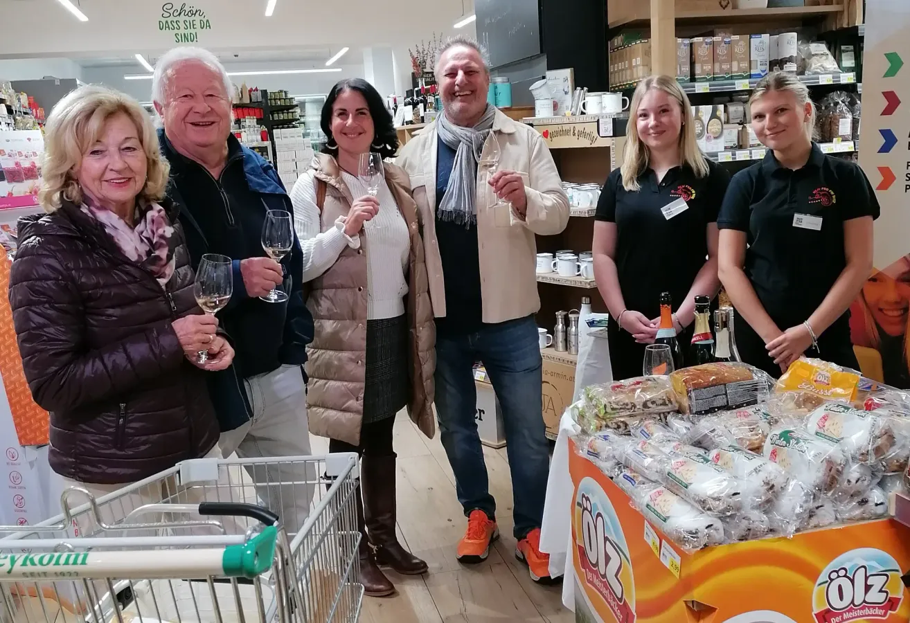 Mehrere Menschen stehen in einem Geschäft, lächeln und halten Weingläser in der Hand, mit einem Tisch voller Lebensmittel vor ihnen. Dahinter sind Regale mit verschiedenen Artikeln gefüllt.