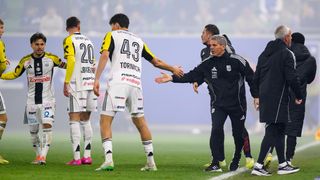 Fußballspieler stehen auf einem Feld. Ein Trainer spricht mit ihnen. Die Spieler tragen Trikots mit Nummern und Namen. Der Hintergrund ist unscharf.