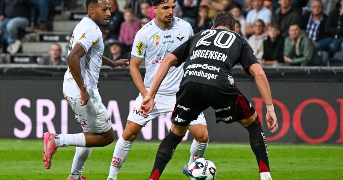 Drei Fußballspieler in Aktion auf einem Feld, einer in Weiß, ein anderer in Weiß mit roten Akzenten und einer in Schwarz mit roten Streifen, sie spielen vor einem verschwommenen Publikum.