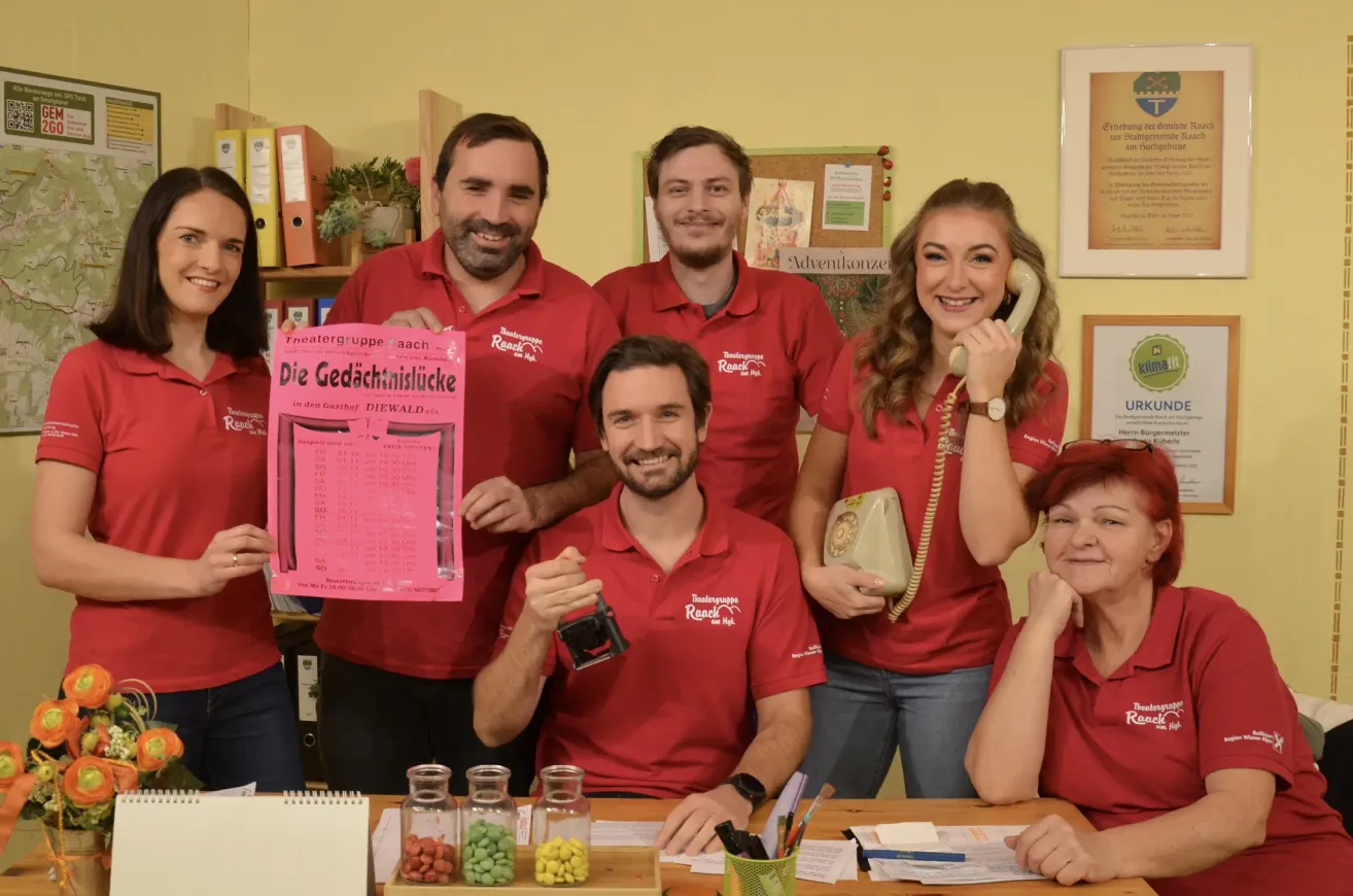 Eine Gruppe von Menschen in roten Poloshirts versammelt sich um einen Tisch und lächelt. Ein Mann in der Mitte hält eine rosa Karte, während eine Frau rechts ein Telefon hält. Sie scheinen sich in einem Studio zu befinden.