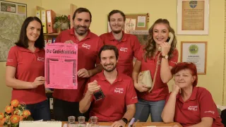 Eine Gruppe von Menschen in roten Poloshirts versammelt sich um einen Tisch und lächelt. Ein Mann in der Mitte hält eine rosa Karte, während eine Frau rechts ein Telefon hält. Sie scheinen sich in einem Studio zu befinden.